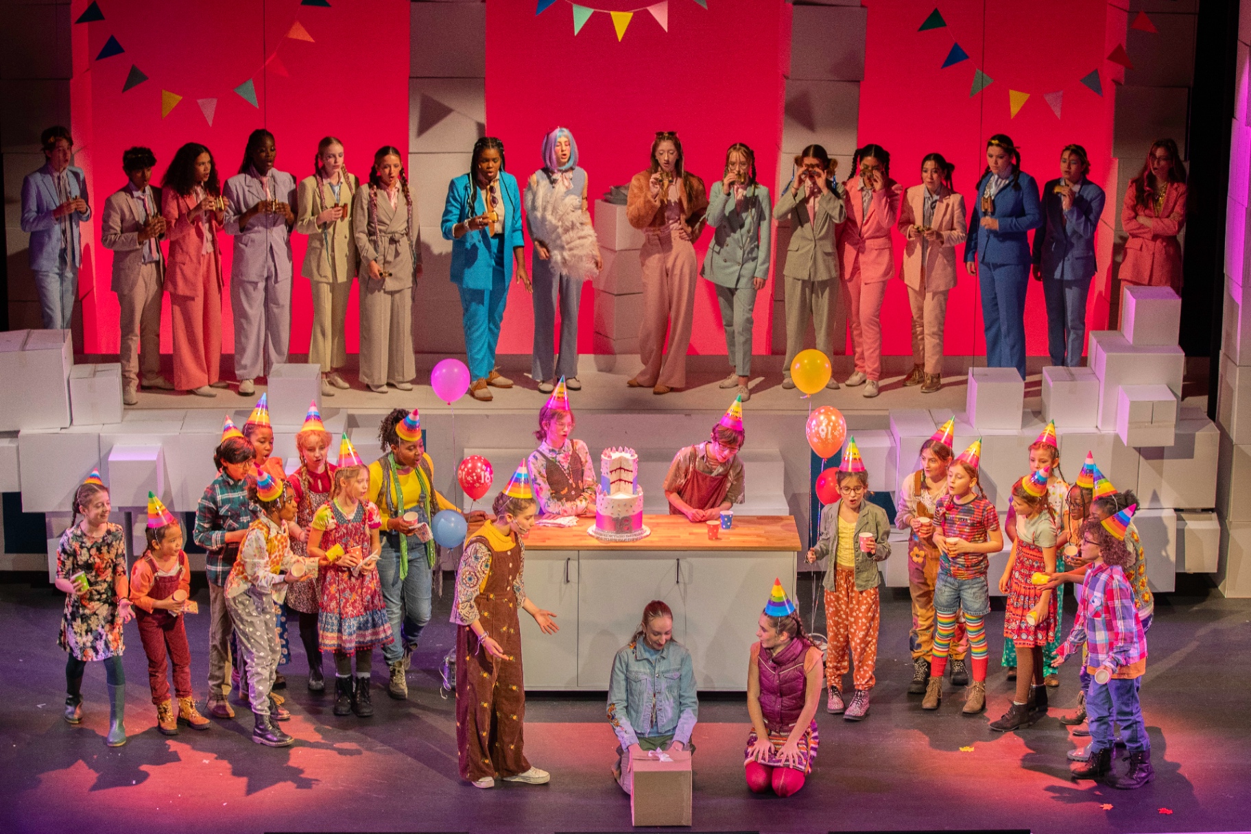 The full company of a London Youth Opera production on stage together — chorus in pastel suits raised above, younger performers in party hats gathered around a birthday cake below.