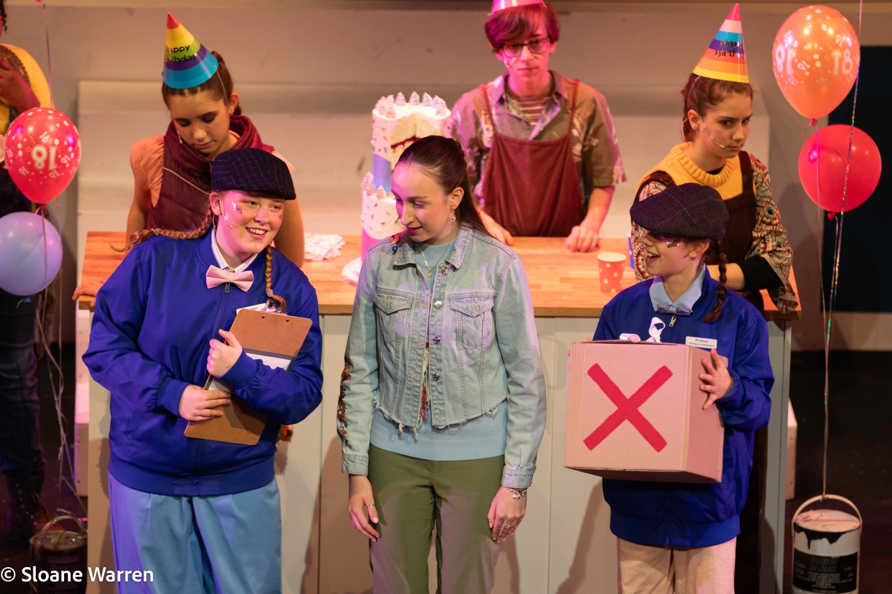Pandora receives a plain cardboard box marked with a red X from two Olympus Enterprises messengers, her family gathered around a birthday cake behind her.