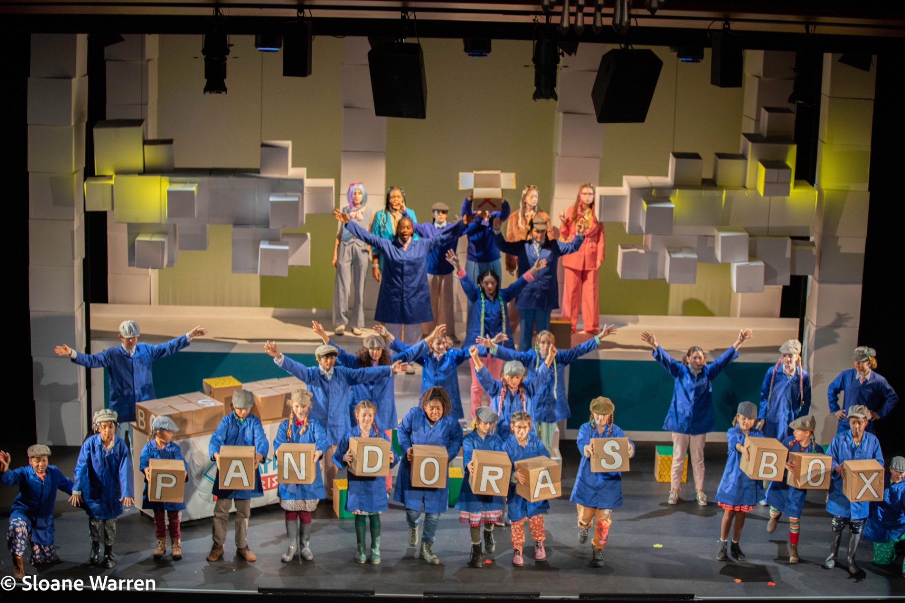 The full company assembled on a set of stacked white cardboard boxes, the mortals in blue overalls and flat caps holding letter blocks spelling 'PANDORA'S BOX' while the gods stand above them.