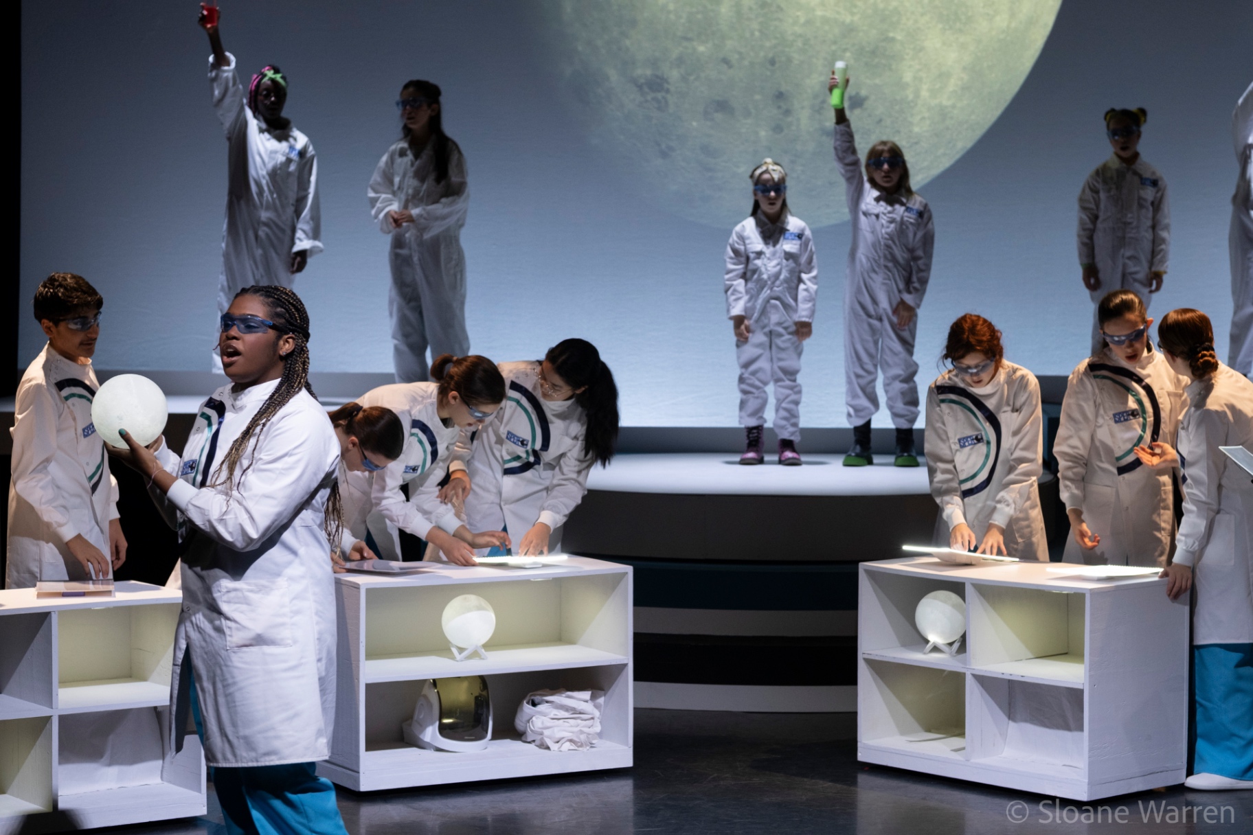The company of young scientists work at illuminated laboratory benches beneath a projected full moon, some standing on a raised platform holding beakers aloft.
