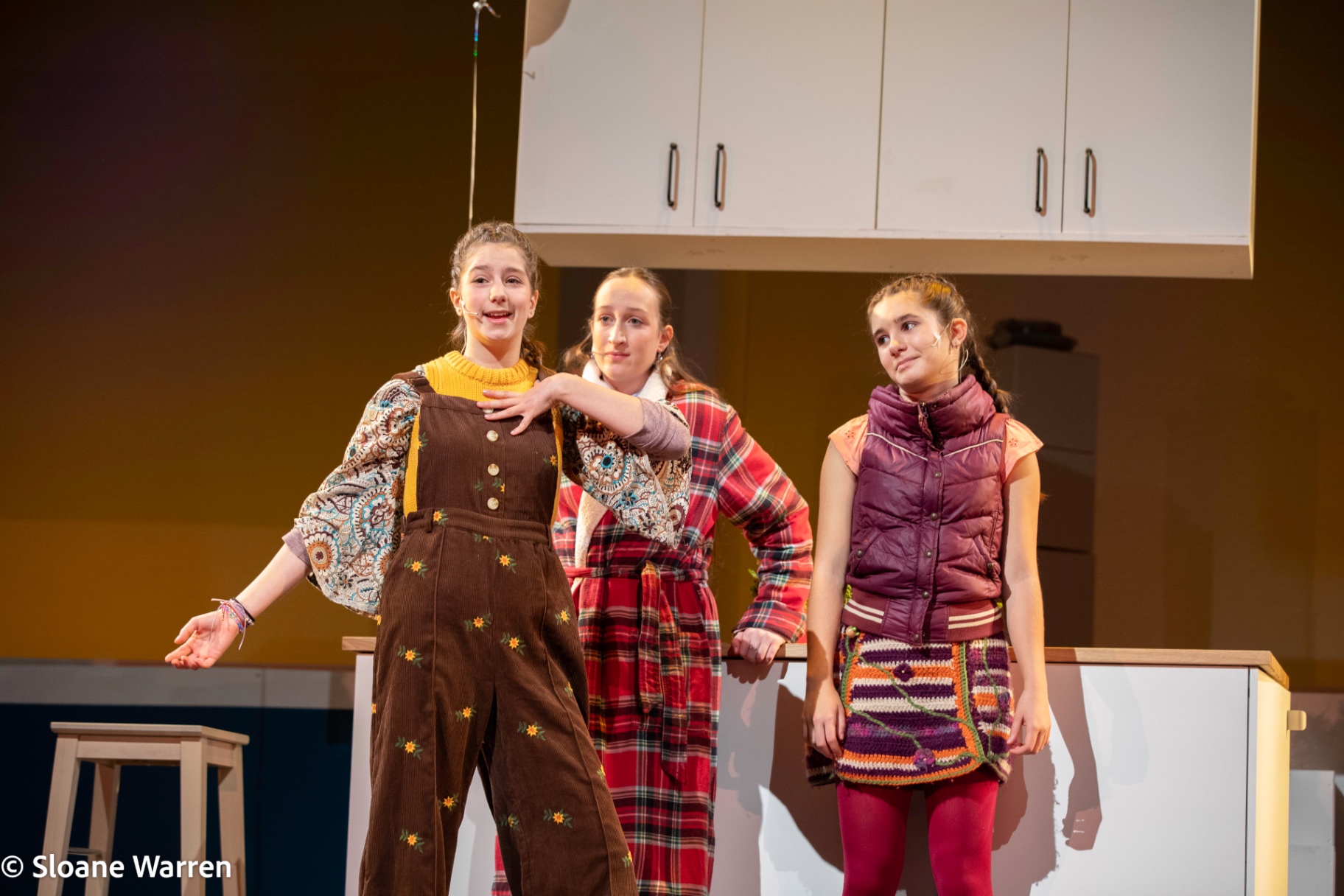 Three friends from the village of Middle Demos gather around a kitchen counter in warm autumnal clothes, mid-conversation.