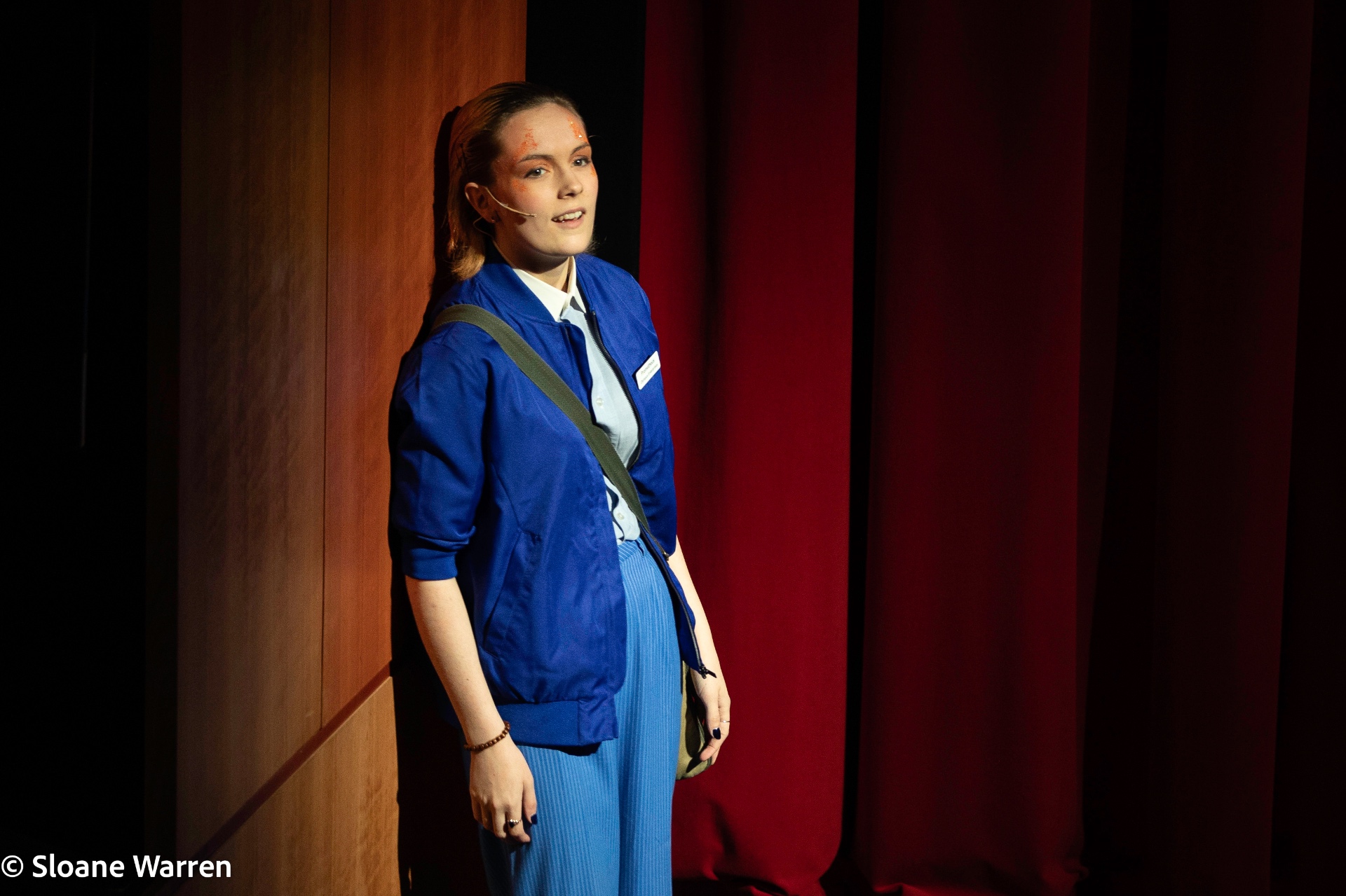 Prometheus, in an Olympus Enterprises blue bomber jacket and messenger bag, stands lit against a red curtain and wooden panelled backdrop.