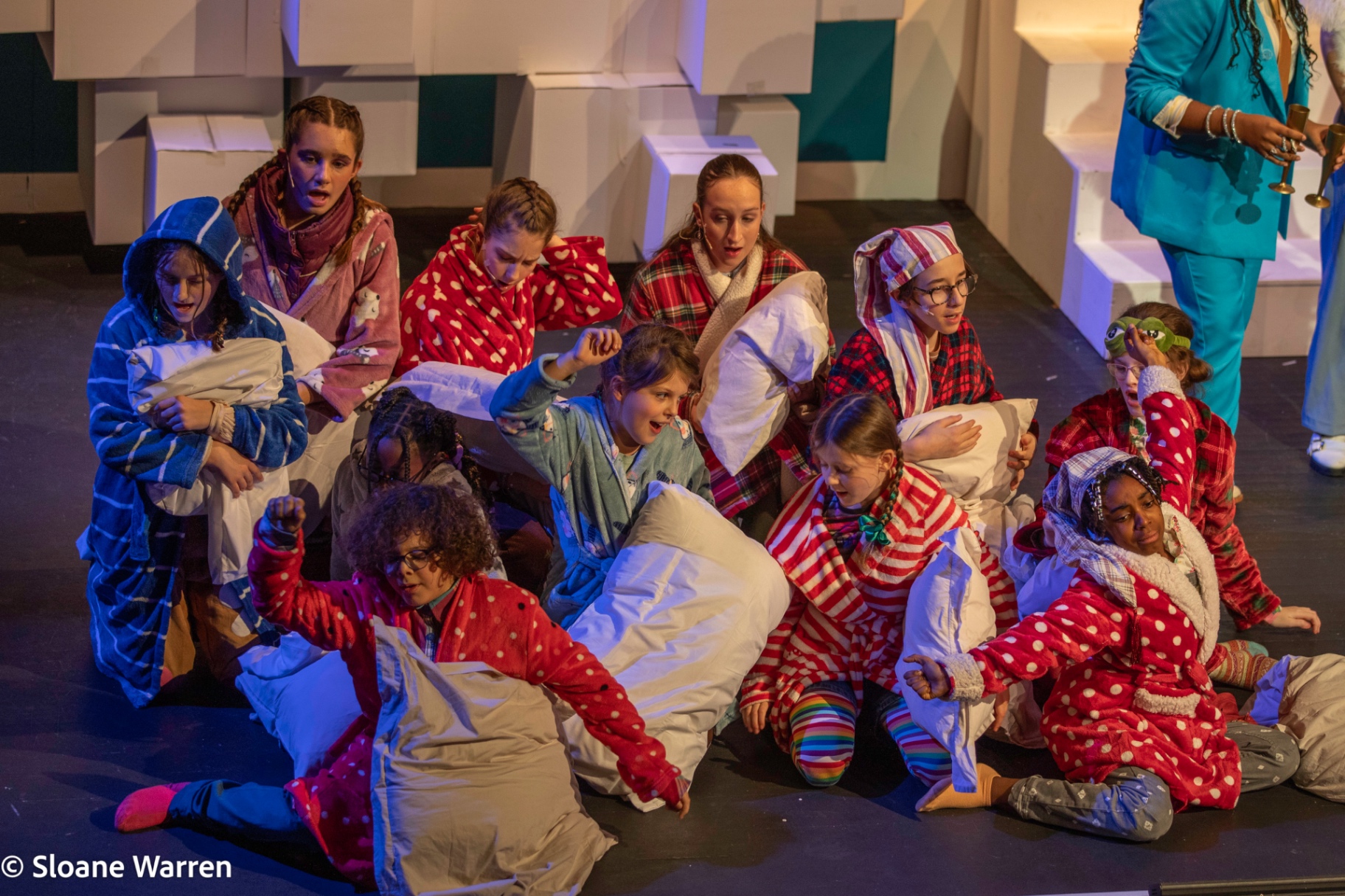 The mortals huddle on the floor in pyjamas and dressing gowns clutching pillows, jolted awake as a god in a turquoise suit stands over them with a trumpet.