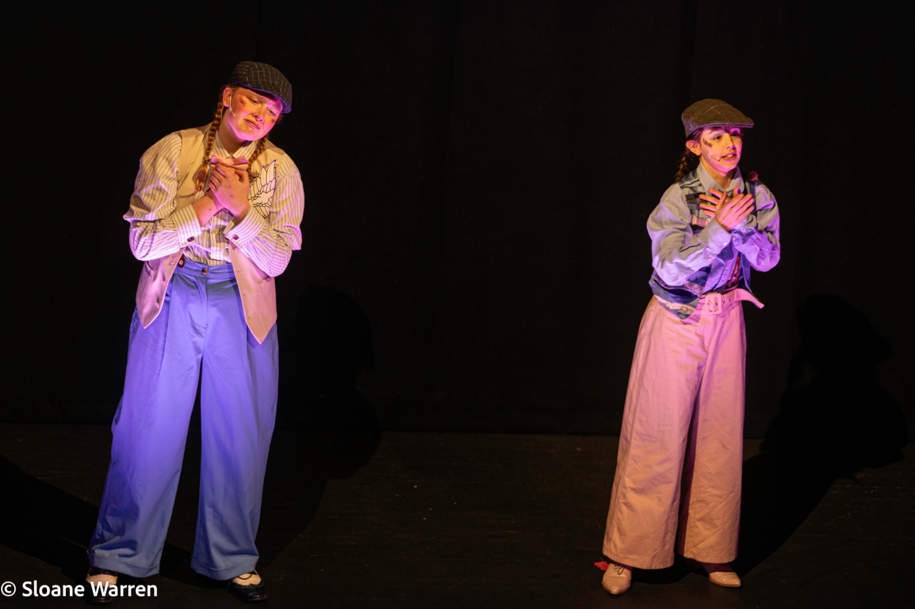 Two Olympus Enterprises messengers in flat caps and wide-legged trousers stand apart on a darkened stage, hands clasped to their chests under warm pink light.